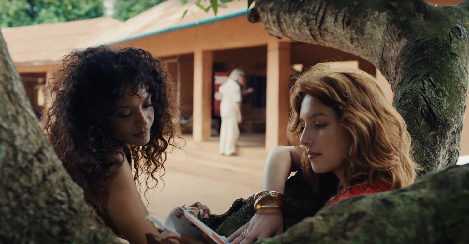 Two travellers reading together under a tree in a red-earth courtyard